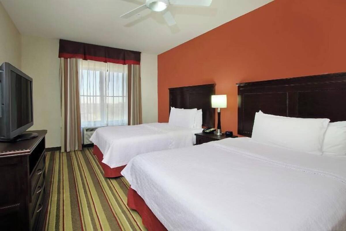 A hotel room with two beds, a television, a ceiling fan, and a window with curtains.