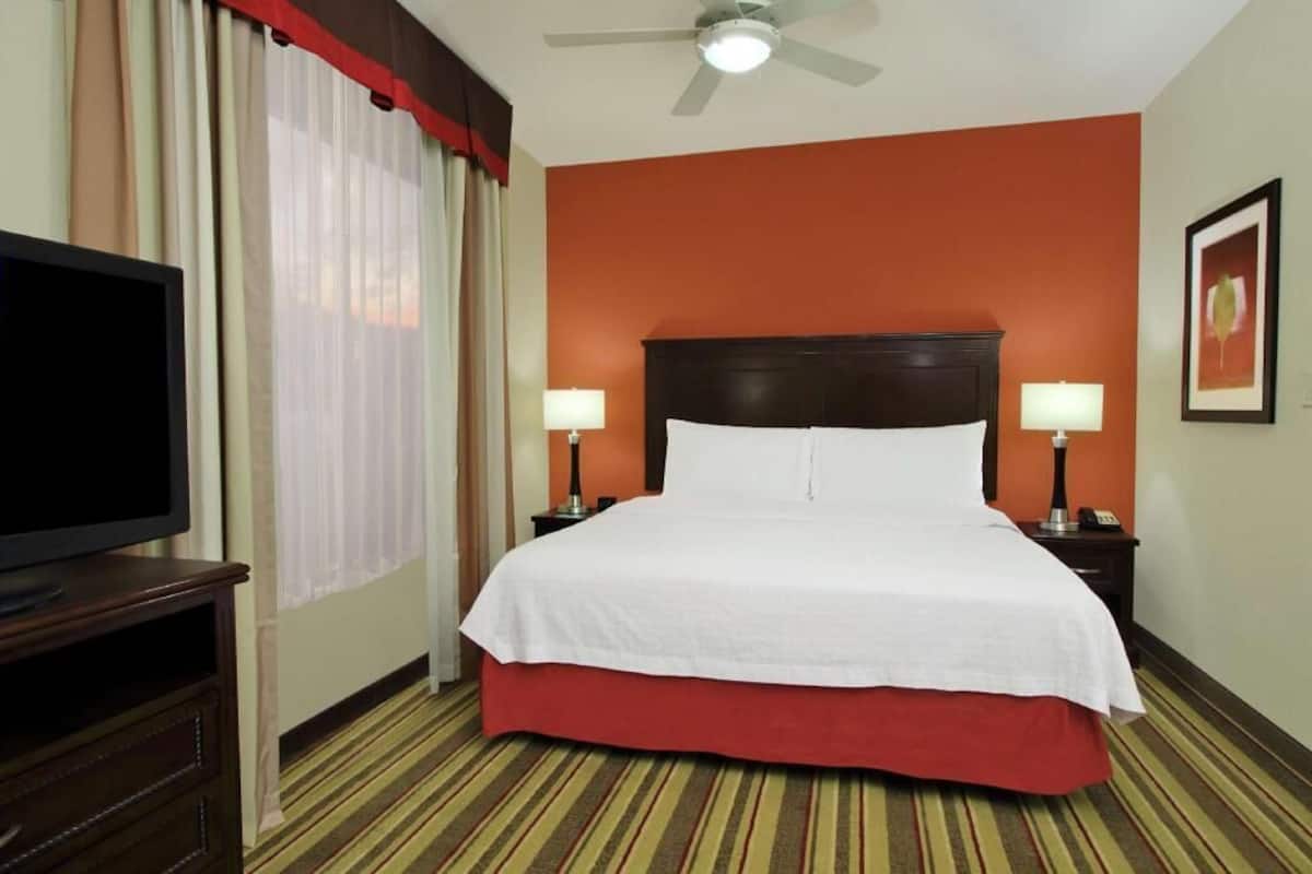 A hotel room with a bed, two bedside lamps, a television on a stand, a ceiling fan, and a window with curtains.