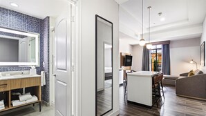 A modern hotel room with a bathroom featuring a large mirror, a vanity with a sink, and a shower. The room has a dining area with a table and chairs, and a living area with a sofa and a TV. - Homewood Beaumont, TX (Beaumont)