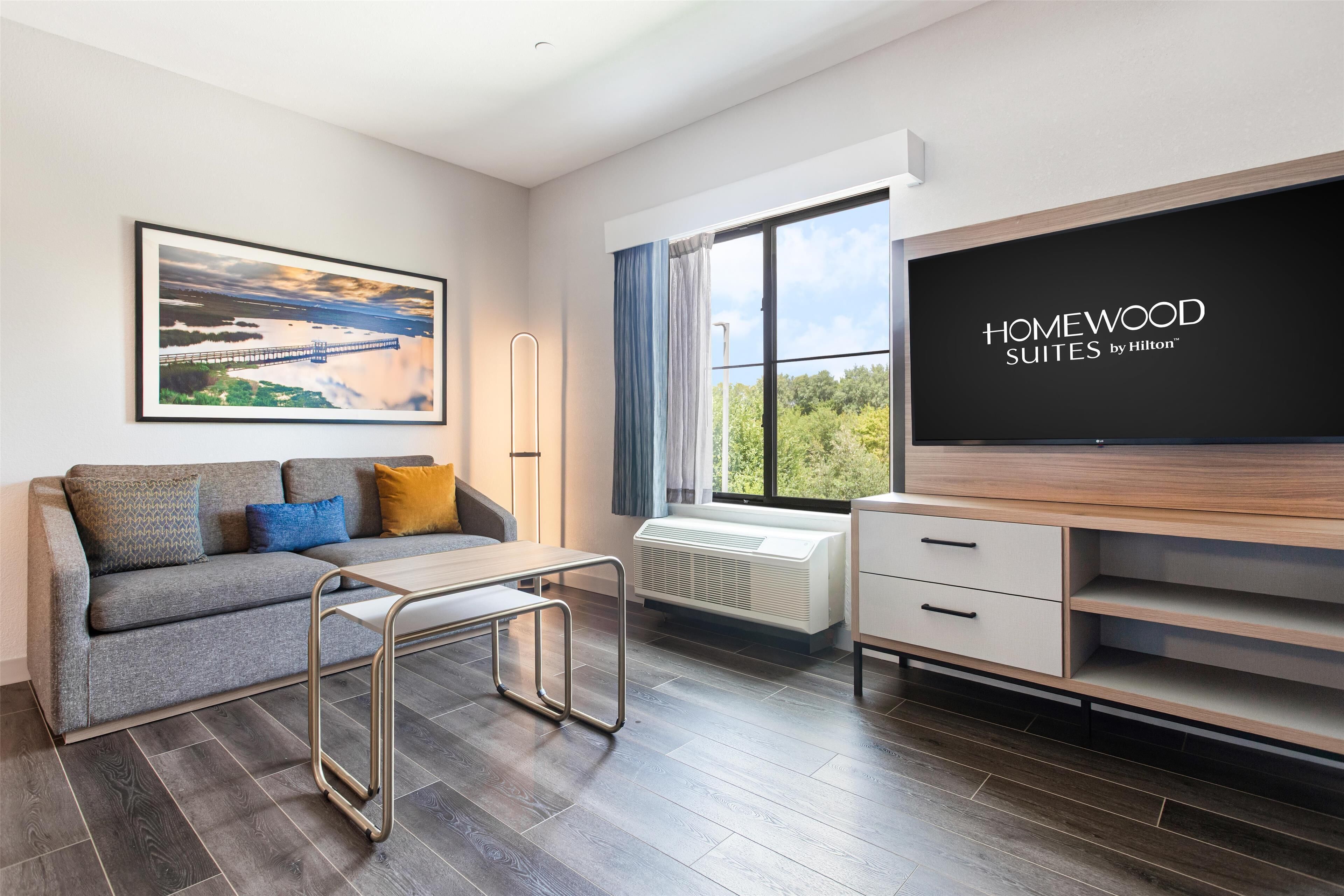 A modern hotel room with a grey sofa, a flat-screen TV, a large window with a view of greenery, and a framed picture on the wall.