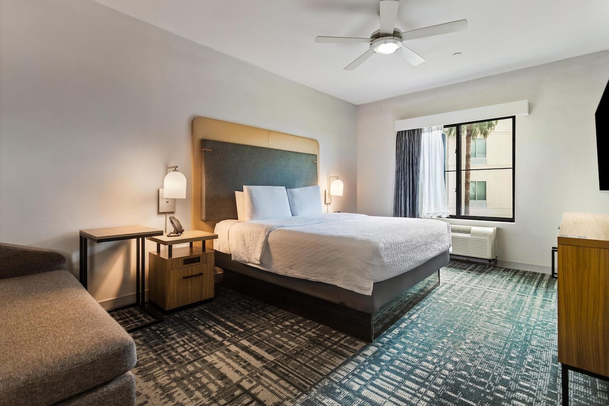 A hotel room with a bed, a desk, a chair, a nightstand, a lamp, a ceiling fan, and a window with curtains.