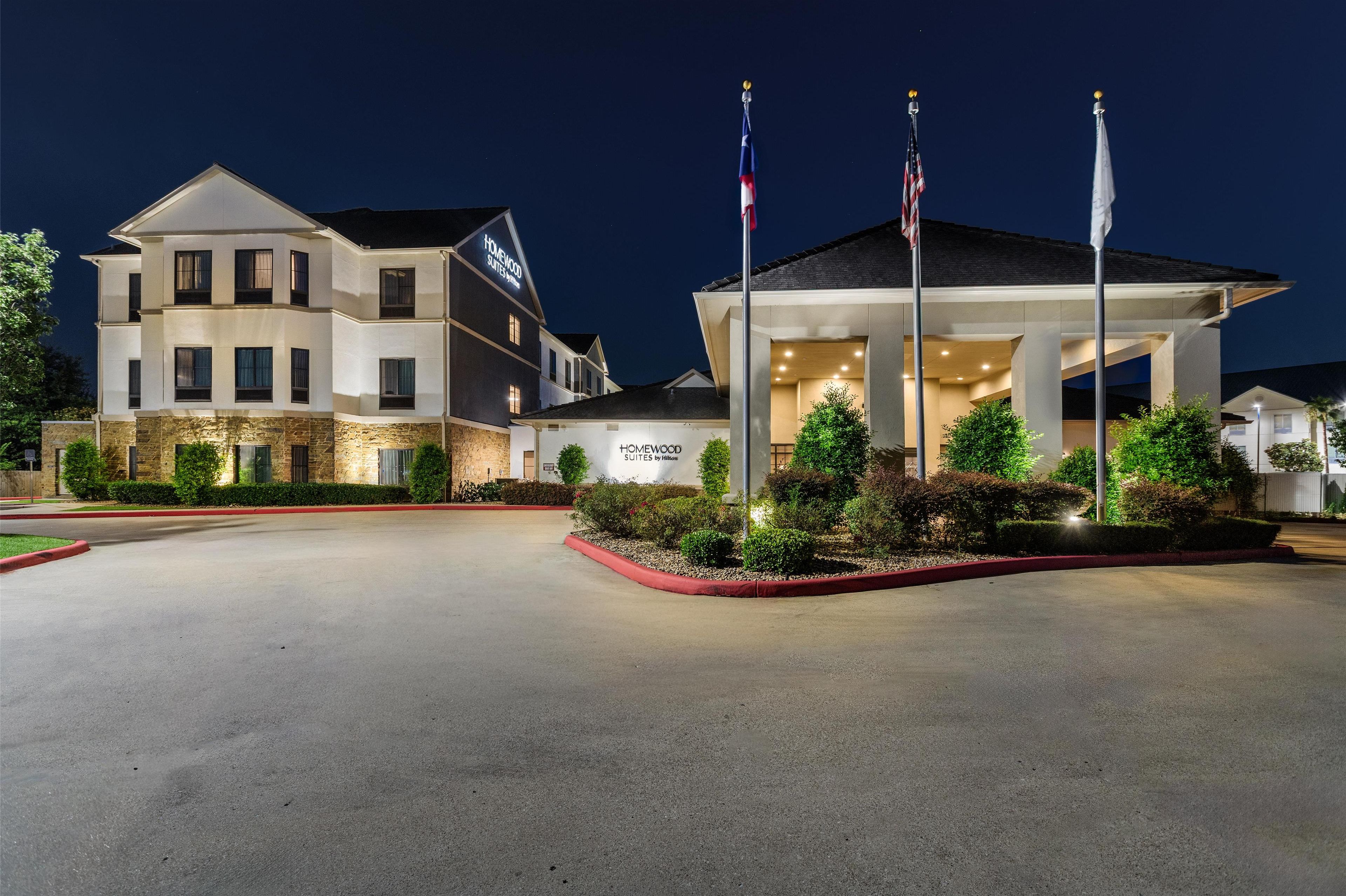 Un hotel de diseño moderno, con una entrada amplia adornada con banderas, fachada bien iluminada y un área ajardinada con arbustos.