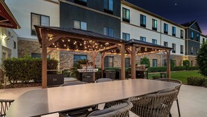 An outdoor dining area with a long table and wicker chairs, a grill, and a covered seating area with string lights. - Homewood Beaumont, TX (Beaumont)