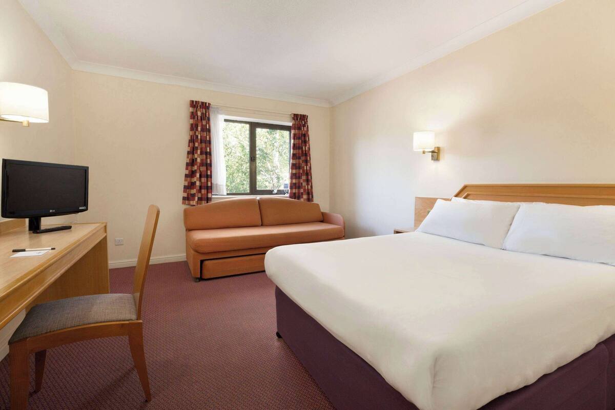 Une chambre d’hôtel comprenant un lit, un bureau avec un ordinateur, une chaise, un canapé et une fenêtre avec des rideaux.