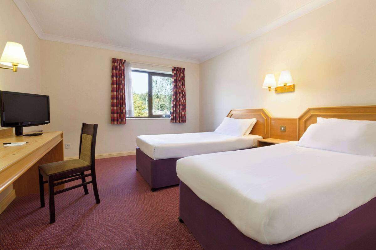 Une chambre d’hôtel avec deux lits simples, un bureau avec une télévision, une chaise et une fenêtre avec des rideaux.