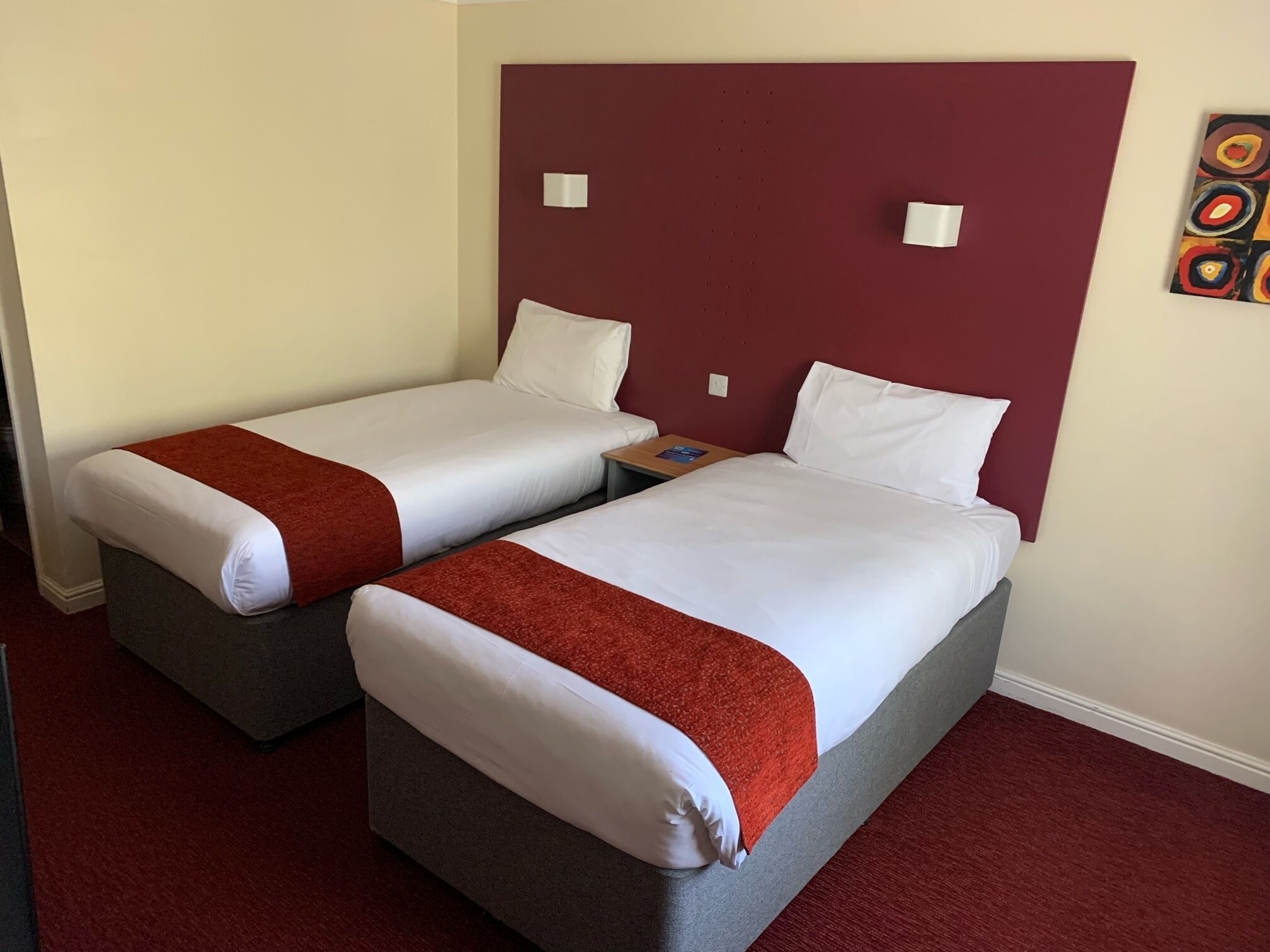 Hotellihuoneessa on kaksi erillistä sänkyä, joissa on valkoiset lakanat ja punainen peitto. Huoneessa on pieni yöpöytä, jossa on lamppu ja kirja. Huoneen sängynpääty on punainen, ja seinällä on taulu.