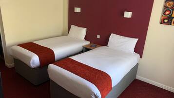Een hotelkamer met twee eenpersoonsbedden, elk met wit beddengoed en een rode deken. Er staat een klein nachtkastje met een lamp en een boek. De kamer heeft een rood hoofdeinde en een schilderij aan de muur.