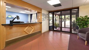 A reception area with a wooden desk, a computer monitor, and a sign for Days Inn. There is a glass door leading outside, and a potted plant is visible. - Days Inn by Wyndham Southampton Rownhams (Southampton)