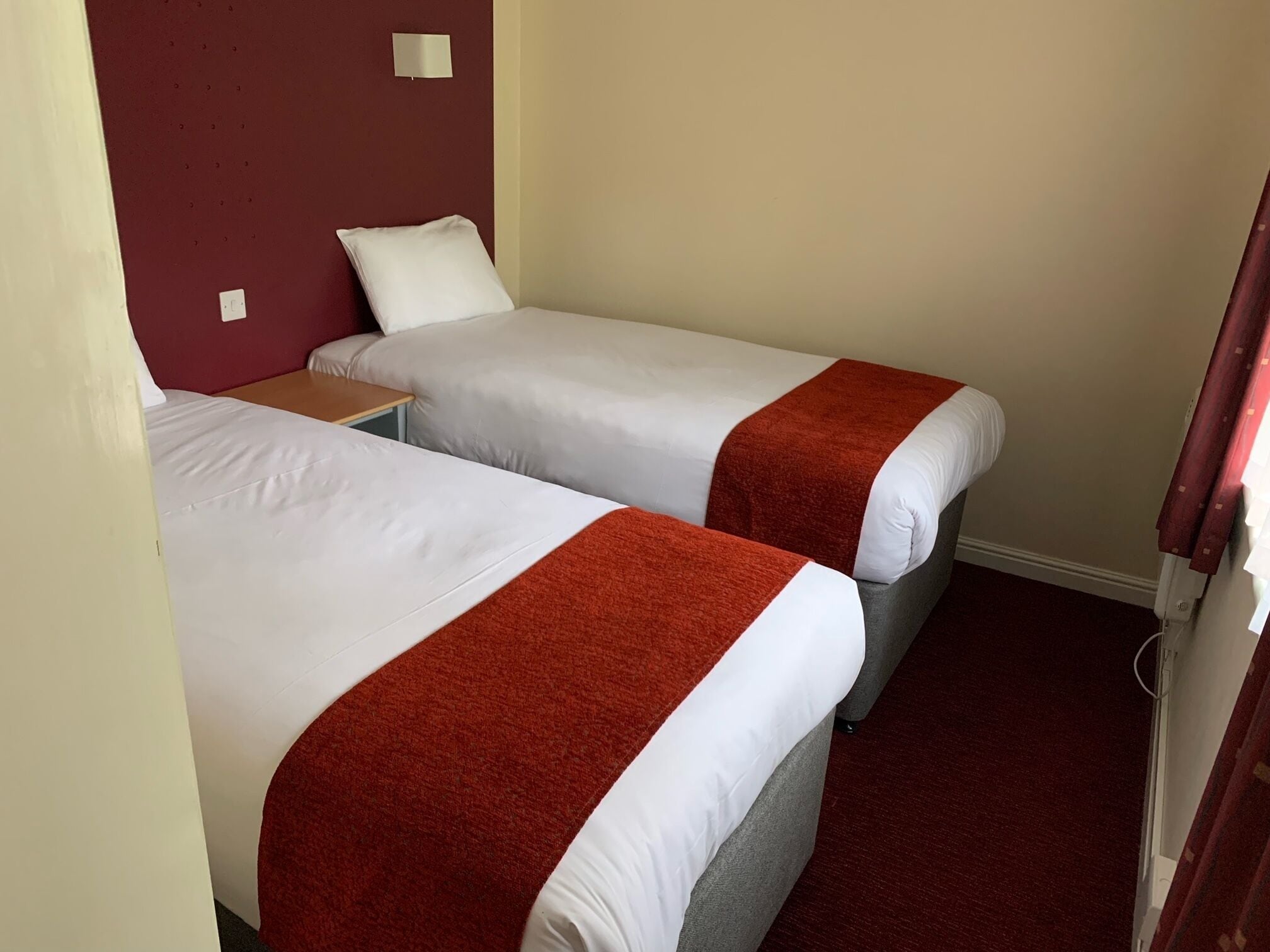 Een hotelkamer met twee enkele bedden, elk met een witte matras en een rode deken. Daartussen staat een klein tafeltje, en erboven een wandlamp.
