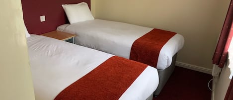 A hotel room with two single beds, each with a white mattress and a red blanket. There is a small table between the beds, and a wall-mounted light fixture above it.