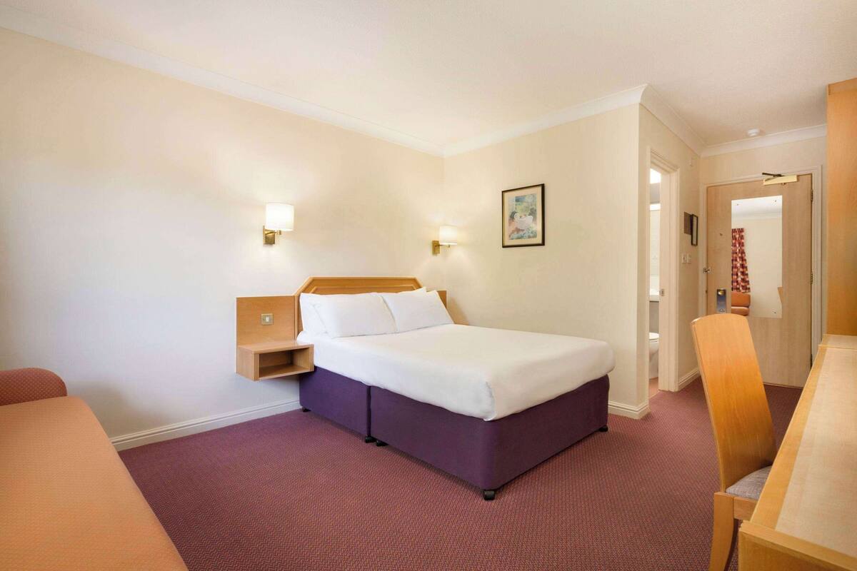 Une chambre d’hôtel avec un lit, un bureau et une chaise. Il y a une table de chevet avec une lampe et un tableau accroché au mur.