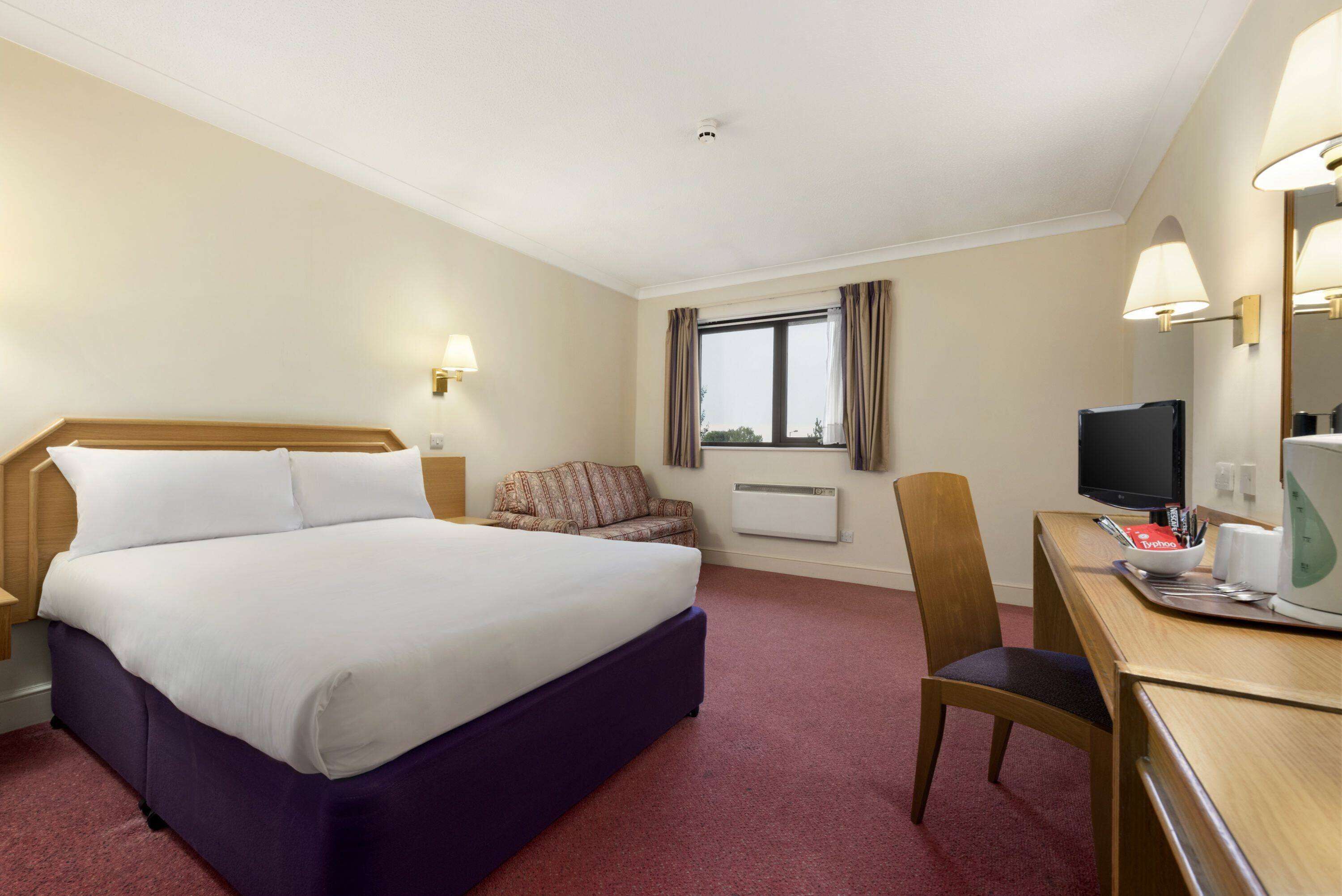 Habitación de hotel con una cama grande, un escritorio con computadora, una silla, una ventana con cortinas y un televisor.