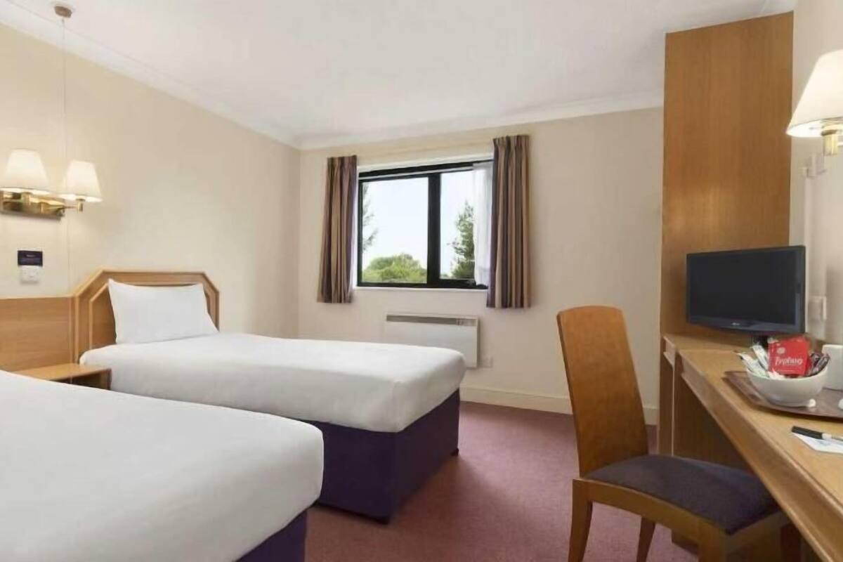 Ett hotellrum med två sängar, ett skrivbord med en stol, en tv och ett fönster med gardiner.
