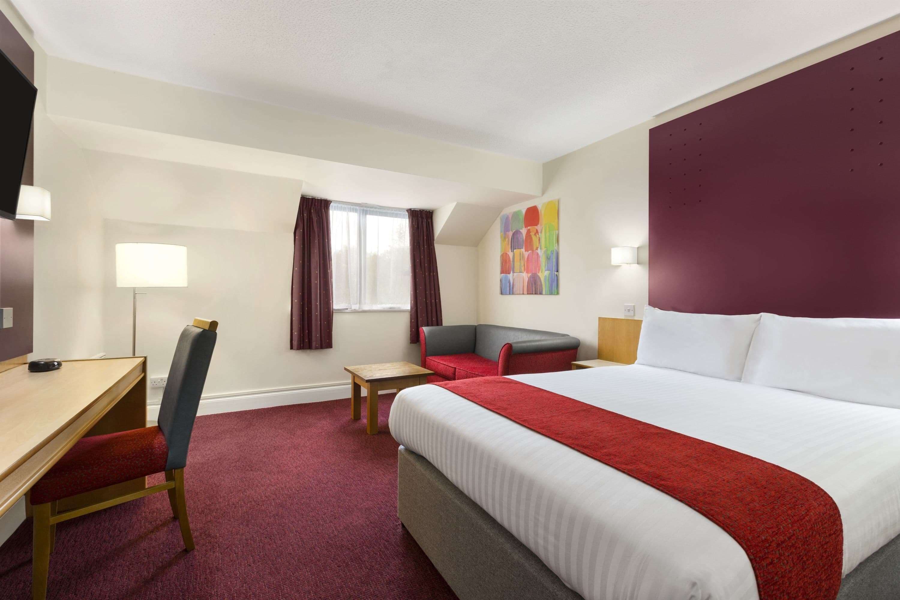 A hotel room with a bed, a desk, a chair, a small table, and a window with curtains.
