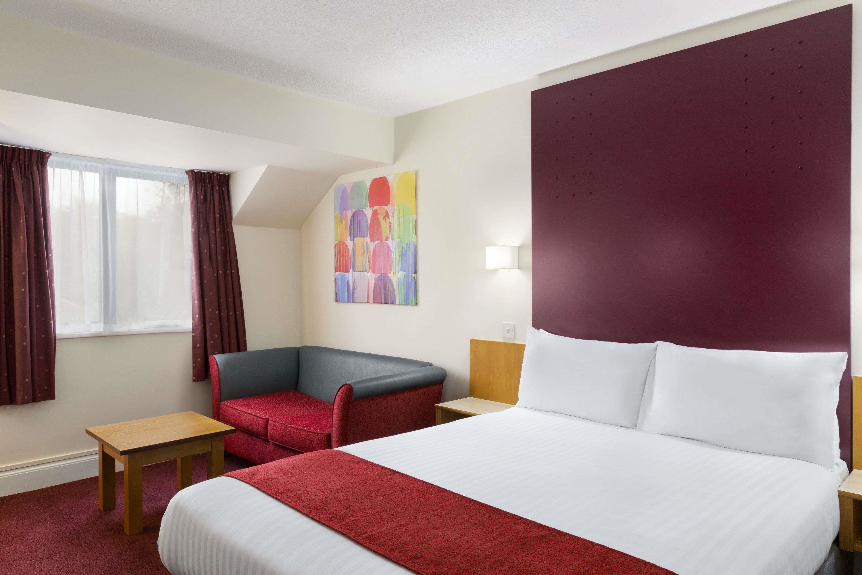 A hotel room with a bed, a red armchair, a small table, and a colorful abstract painting on the wall.