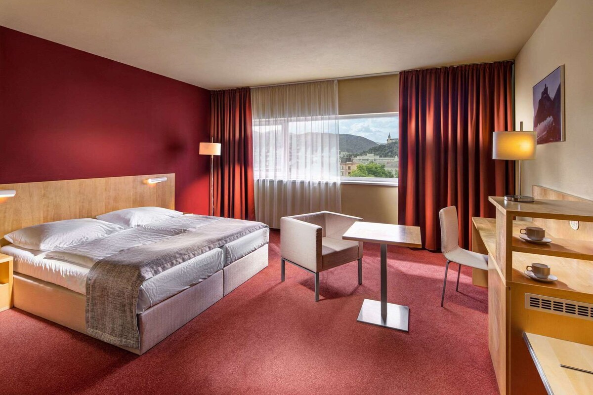 A hotel room with a bed, a desk, a chair, a table, and a view of the city.