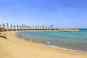 A beach with clear blue water, palm trees, and thatched-roof structures along the shoreline. - Meraki Resort - Adults Only - All inclusive (Hurghada)