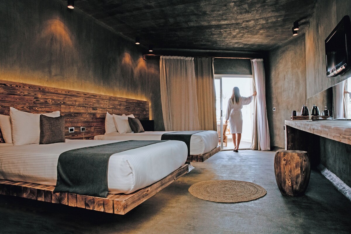 Una habitación de hotel moderna con dos camas, un cabecero de madera, un taburete de madera redondo y un gran ventanal con cortinas.