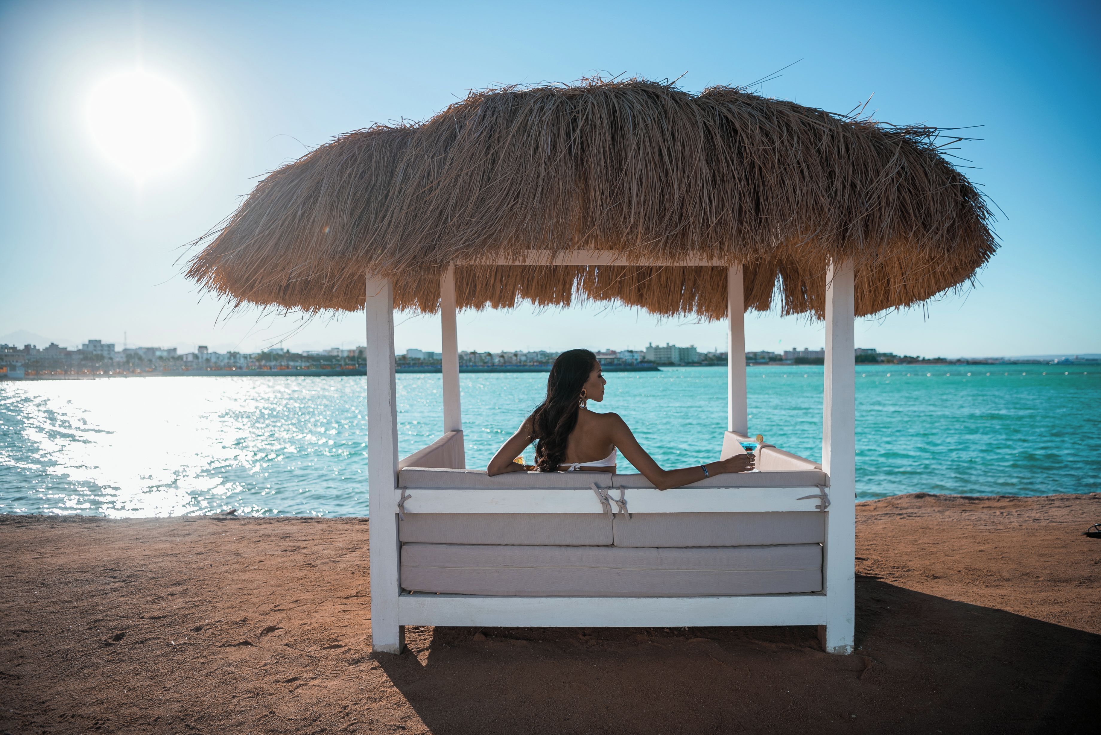 A beachside cabana with a thatched roof, a person relaxing, and a view of the sea and distant buildings.