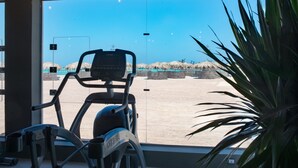 A stationary bike in a room with a large window overlooking a beach with thatched umbrellas. - Meraki Resort - Adults Only - All inclusive (Hurghada)