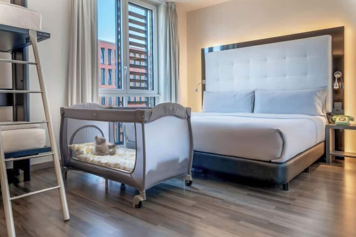 Una habitación de hotel con una cama, una cuna, una escalera y una ventana con vistas a edificios.