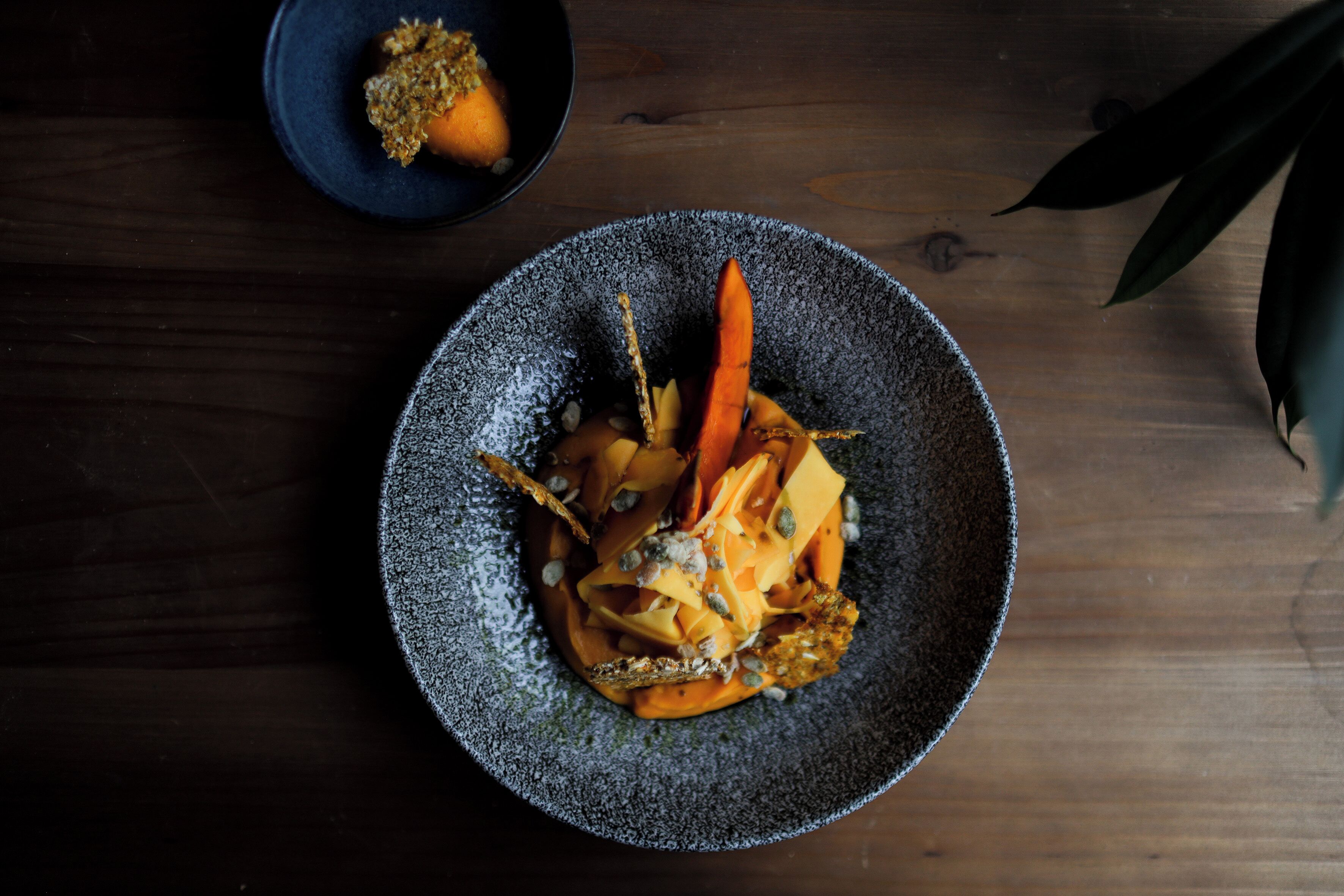 A dark wooden table with a textured bowl containing a dish with vegetables and a carrot.