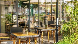 Ein Straßencafé mit Tischen und Stühlen, umgeben von Glaswänden und Pflanzen.