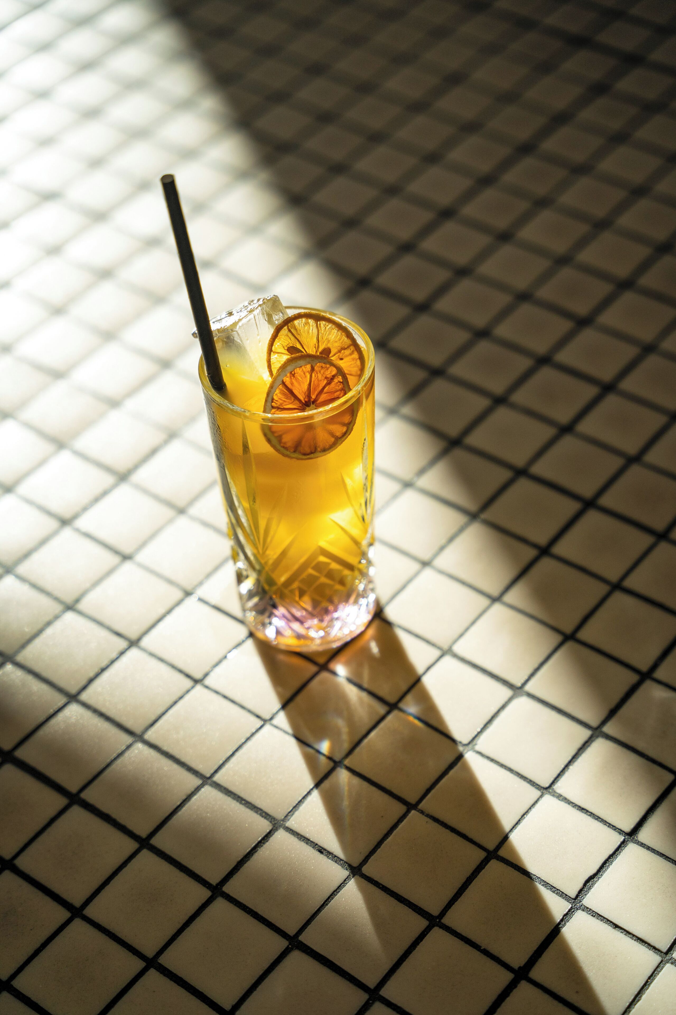 Un vaso de bebida color ámbar con pajita y una rodaja de naranja sobre una superficie de baldosas.