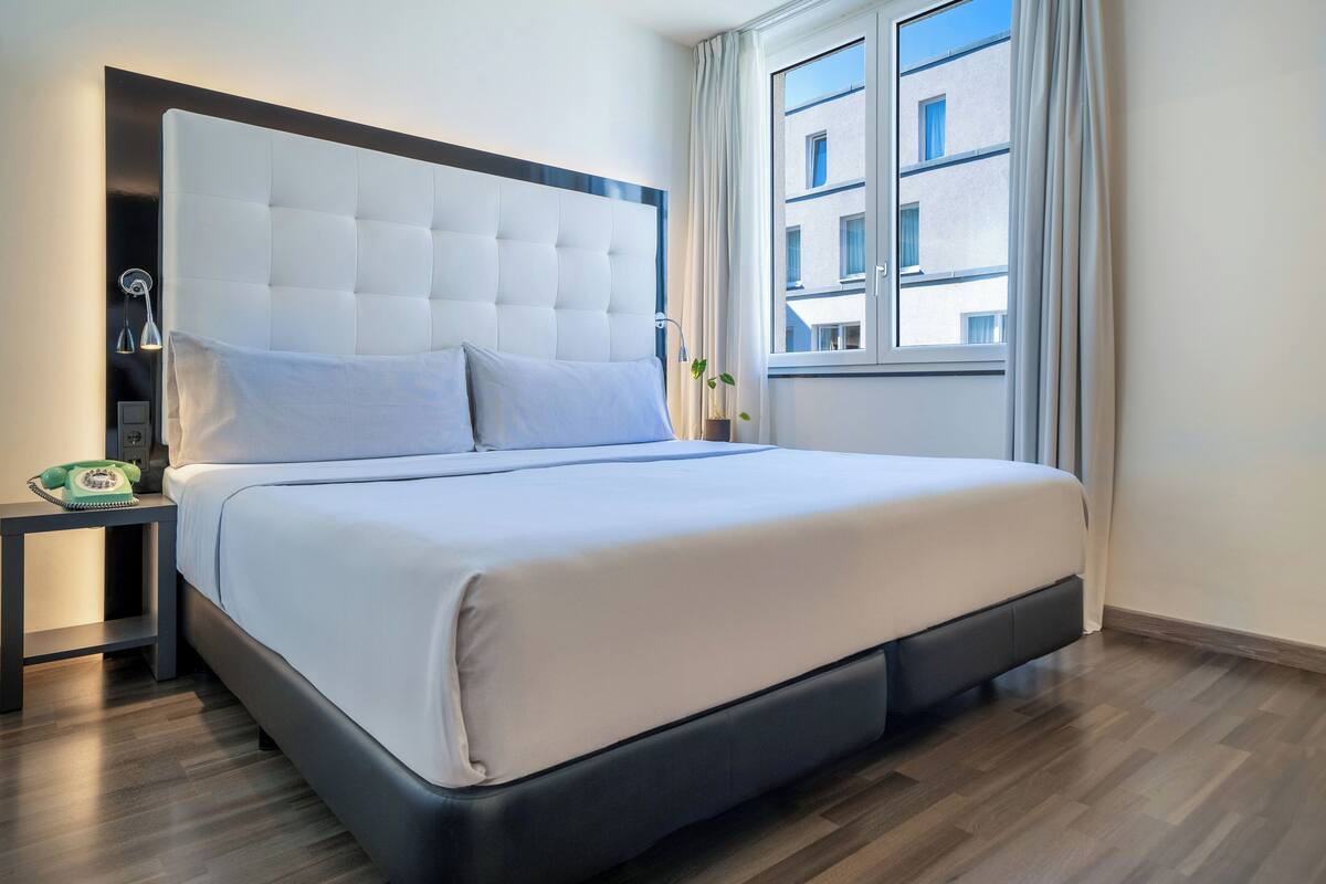 Una habitación de hotel moderna con una cama grande, una mesita de noche con lámpara y vistas a la ciudad desde la ventana.