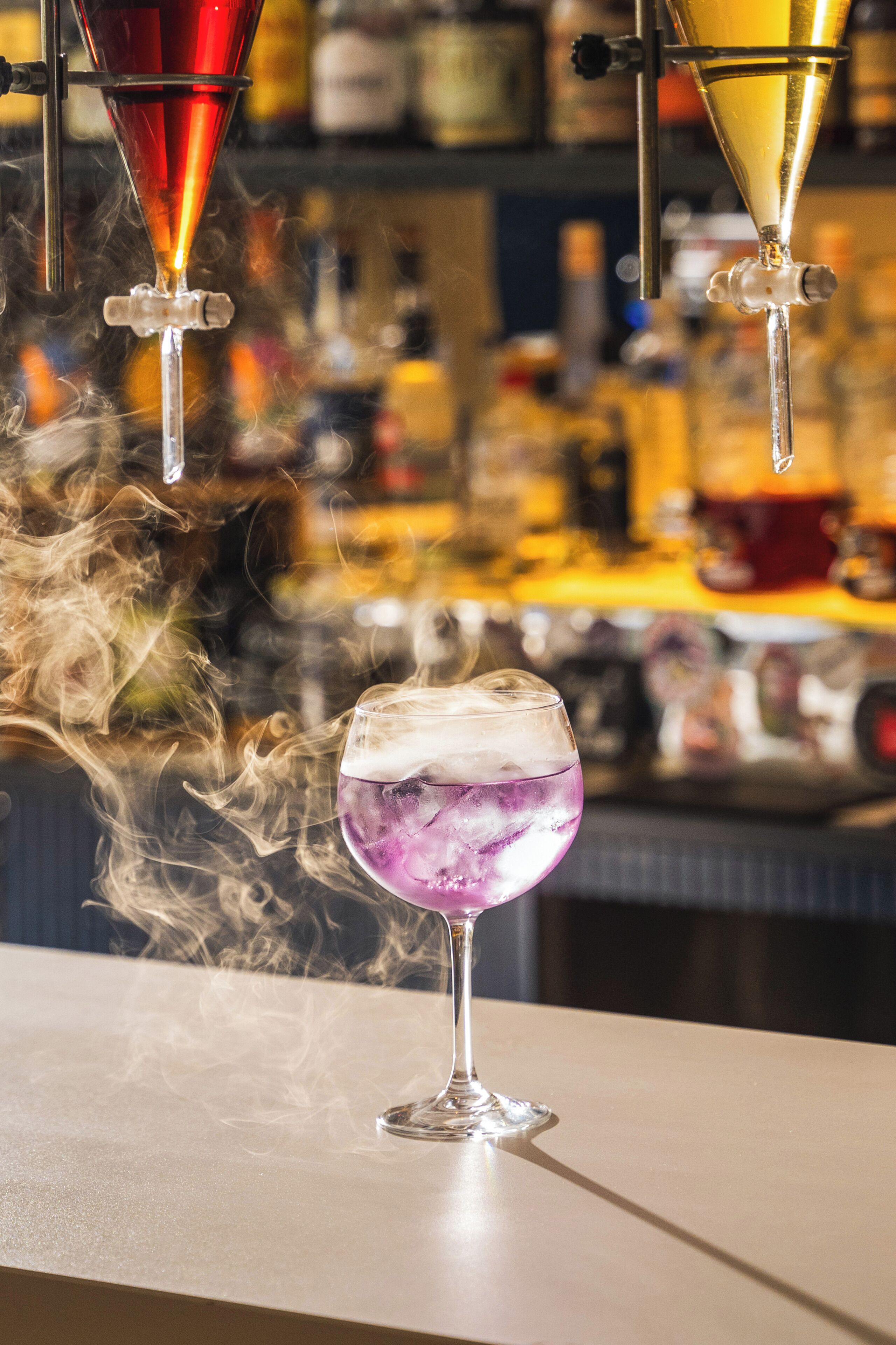 A bar setting with a drink in a glass, steam rising from the drink, and a blurred background of bottles and bar equipment.