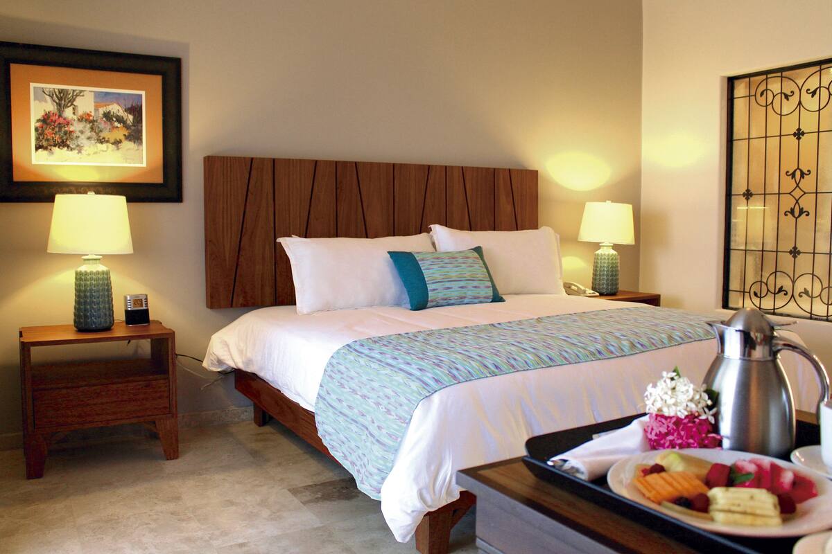 A hotel room with a wooden headboard, two bedside tables with lamps, a bed with white bedding and a patterned throw, and a tray with fruits and a coffee pot.