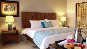 Habitación de hotel con cabecera de madera, dos mesitas de noche con lámparas, cama con ropa blanca y un plaid estampado, y una bandeja con frutas y una cafetera.