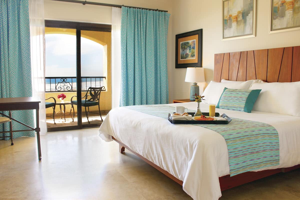 A hotel room with a bed, a desk, a chair, a balcony with a view, and a table with a tray of breakfast items.