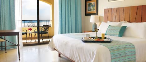 Habitación de hotel con cama, escritorio, silla, balcón con vistas y una mesa con una bandeja de productos para el desayuno.