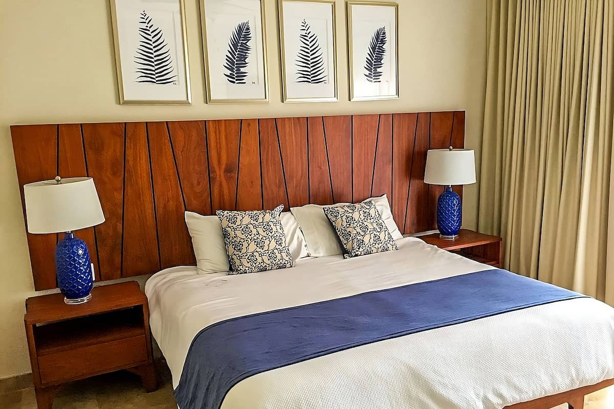 A hotel room with a wooden headboard, a large bed with a blue and white bedspread, two nightstands with lamps, and four framed leaf artworks on the wall.