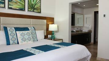 A hotel room with a large bed, a nightstand with a lamp, a bathroom with a sink and mirror, and artwork on the wall.