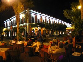 Un espacio de comedor al aire libre con mesas y sillas, una palmera, y un edificio con ventanas iluminadas y columnas.