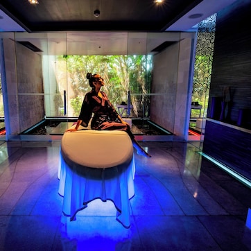A modern spa room with a massage table, glass walls, and a view of greenery outside.