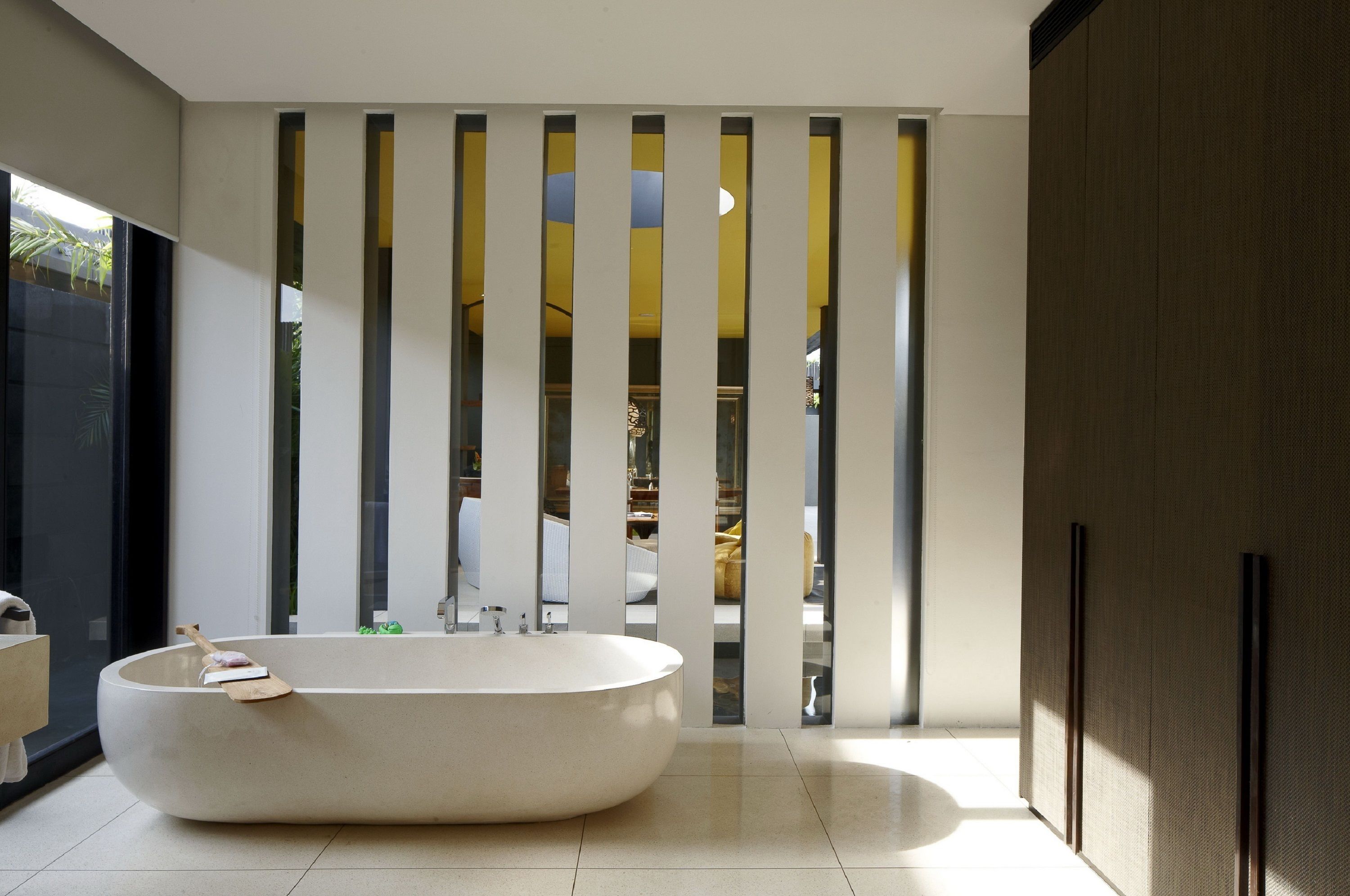 A modern bathroom with a freestanding bathtub, a large window, and a view of a room with yellow furniture.