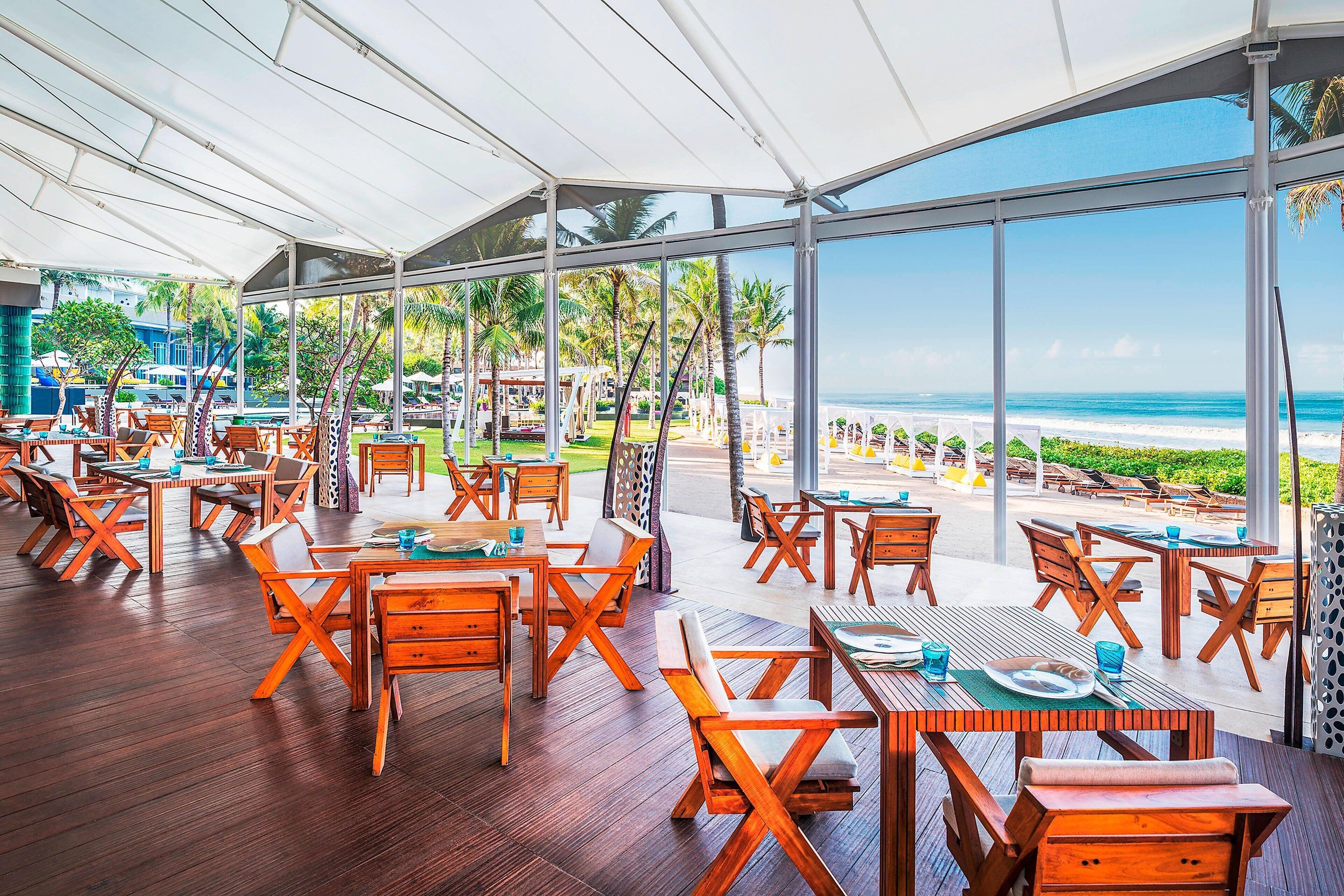 A covered outdoor dining area with wooden tables and chairs, overlooking a beach.