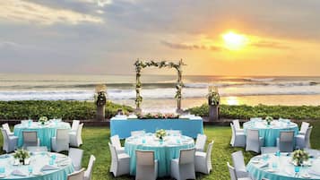 Une cérémonie de mariage en plein air avec des tables rondes, des chaises blanches et une arche fleurie surplombant l’océan.