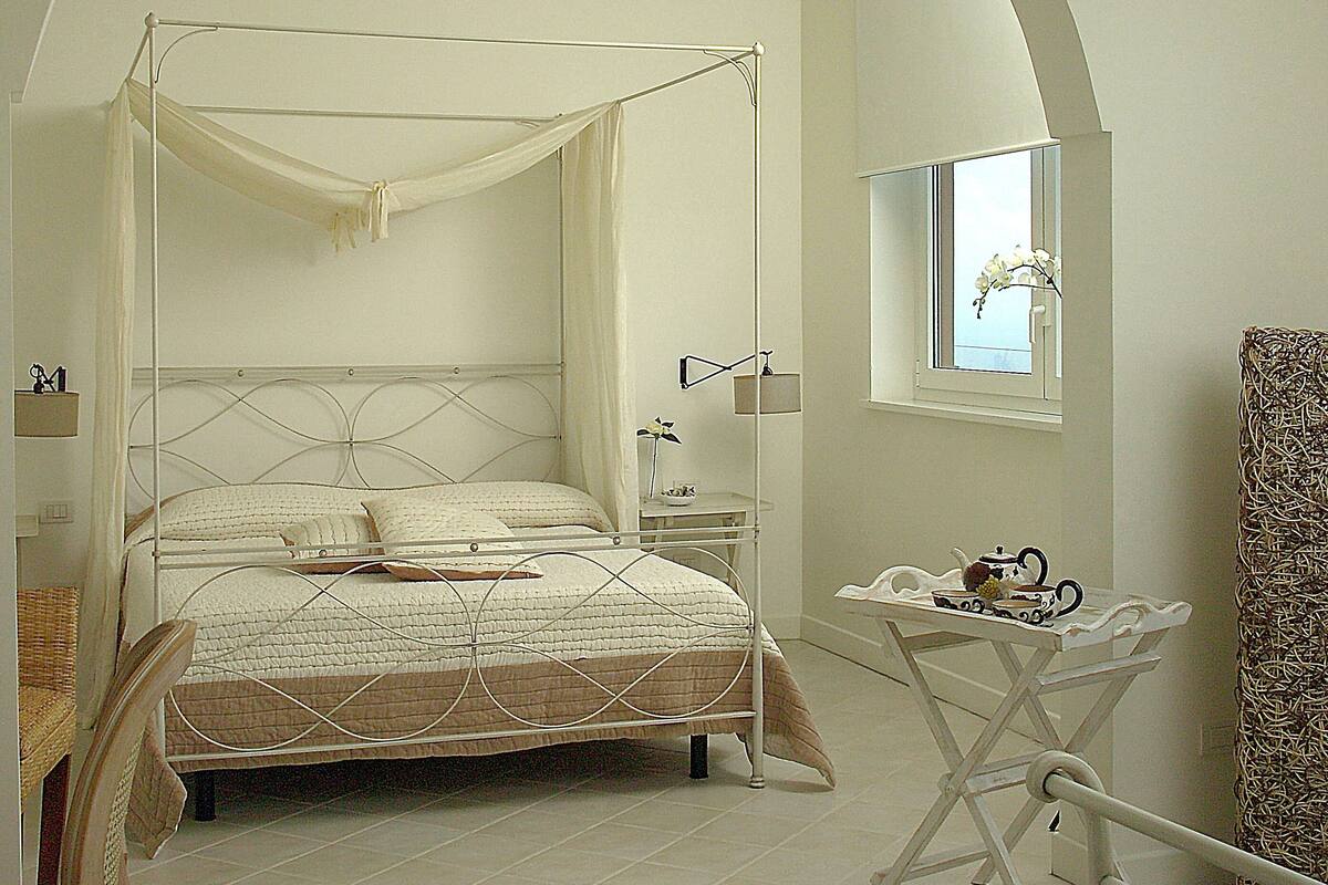 Een slaapkamer met een hemelbed, een klein tafeltje met een theepot en een raam met uitzicht op de lucht.