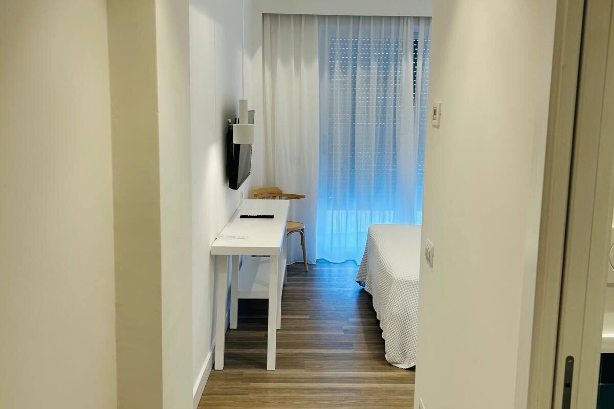 Een smalle gang die uitkomt in een slaapkamer met een bed, een bureau en een stoel.