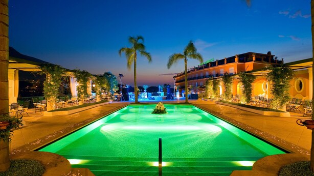 Vista nocturna de un área de piscina de hotel con una fuente central, palmeras y asientos al aire libre.