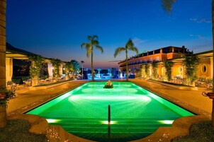 Vista nocturna de un área de piscina de hotel con una fuente central, palmeras y asientos al aire libre.