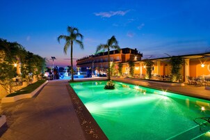 Vista nocturna de un hotel con piscina, palmeras y zonas de estar al aire libre.