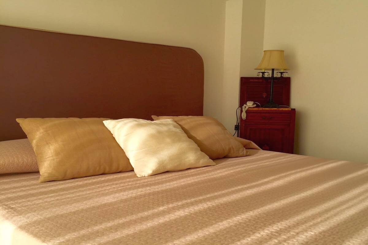 Een keurig opgemaakt bed met een gestreept dekbed en twee kussens, een houten nachtkastje met een lamp en een telefoon.