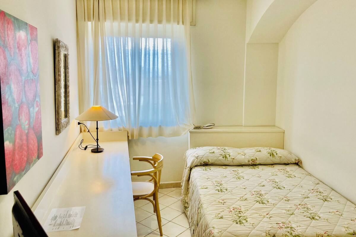 Een kleine, goed verlichte kamer met een bed, een bureau met een lamp, een stoel en een raam met gordijnen.