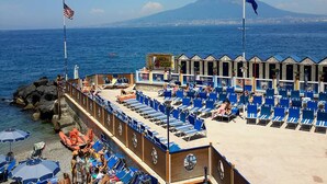 Un hotel frente a la playa con hileras de sillas azules y sombrillas, una costa rocosa y una montaña al fondo.