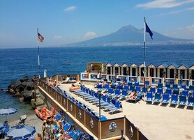 Un hotel frente a la playa con hileras de sillas azules y sombrillas, una costa rocosa y una montaña al fondo.